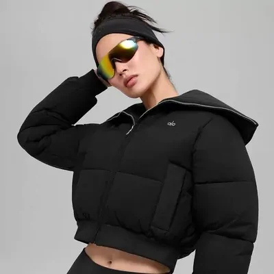 A model in a black cropped puffer jacket and mirrored sunglasses, hand raised to a black headband